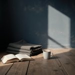 Sala vacía al amanecer con pared índigo, un libro abierto sobre el suelo de madera, una manta gris doblada y una taza de té fría junto a la ventana.